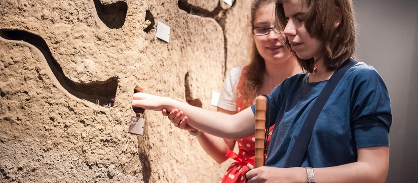 Ein Mädchen mit einer Sehbehinderung betastet ein lebensgroßes Modell aus Stein.Ein anderes Mädchen führt ihr lächelnd den rechten Arm.