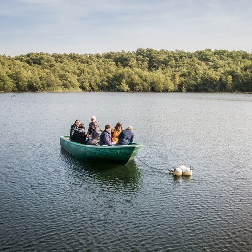 Bick auf See mit Ruderboot und Menschen darin. Im Hintergrund dichter Laubwald.