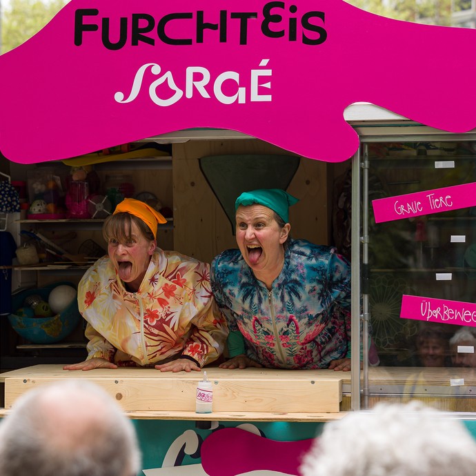 Zwei bunt verkleidete Frauen lehnen sich aus einem Eiswagen und strecken die Zungen heraus. (vergrößerte Bildansicht wird geöffnet)