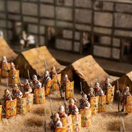 Miniaturfiguren in römischer Rüstung mit bemalten Schilden stehen vor Zelten auf sandigem Untergrund, im Hintergrund ein Modellgebäude mit Fachwerkstruktur.