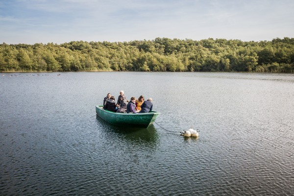 Bick auf See mit Ruderboot und Menschen darin. Im Hintergrund dichter Laubwald.