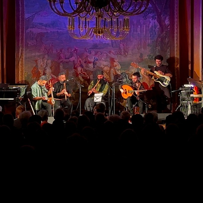 Eine siebenköpfige Band spielt auf einer großen Bühne vor vollem Saal. (vergrößerte Bildansicht wird geöffnet)