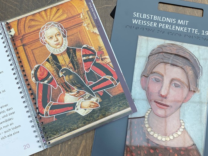 Ein aufgeschlagenes Buch mit Texten in Brailleschrift und Bildern zum Tasten sowie eine Tafel mit einem Gemälde mit Reliefstruktur zum Ertasten. Ein aufgeschlagenes Buch mit Texten in Brailleschrift und Bildern zum Tasten sowie eine Tafel mit einem Gemälde mit Reliefstruktur zum Ertasten.