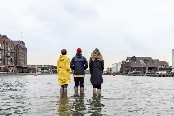 Rückseitige Ansicht dreier Personen, die mitten im Kanal in Münster mit hochgekrempelten Hosen bis zu den Waden im Wasser stehen.