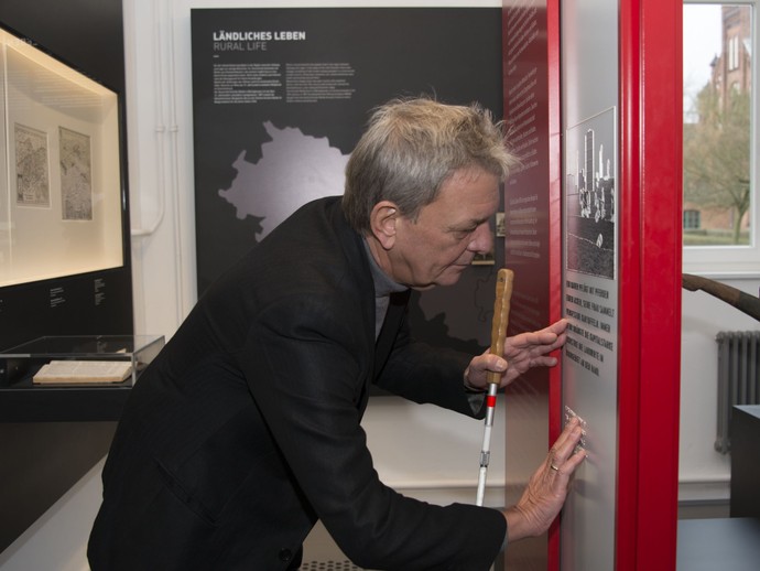Ein Mann mit Blindenstock ertastet einen Informationstext in Braille-Schrift. Ein Mann mit Blindenstock ertastet einen Informationstext in Braille-Schrift.