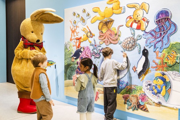 Kinder betrachten eine bunte Wandmalerei mit Meeresbewohnern und Hasenfiguren, daneben steht eine Person im großen Hasenkostüm mit rotem Halstuch und roten Schuhen