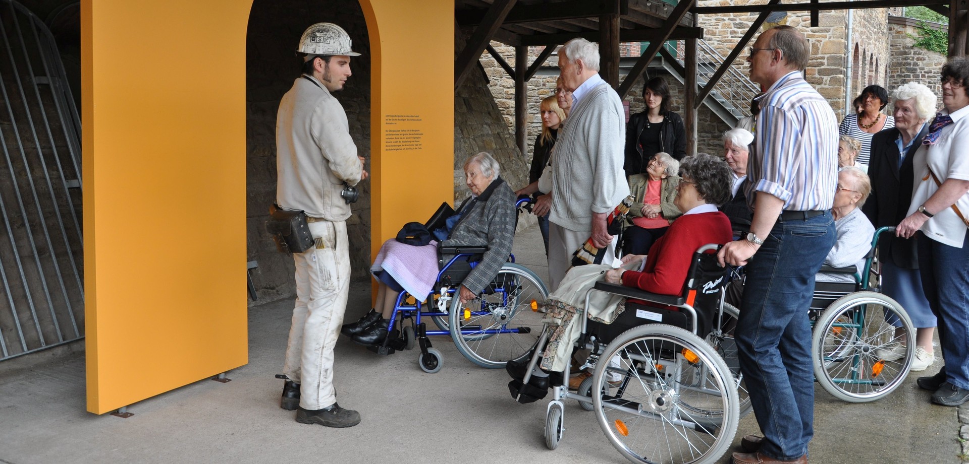 Mehrere Personen im Rollstuhl, stehende Personen und ein Museumsführer haben sich um den Eingang des Bergwerks der Zeche Nachtigall versammelt.