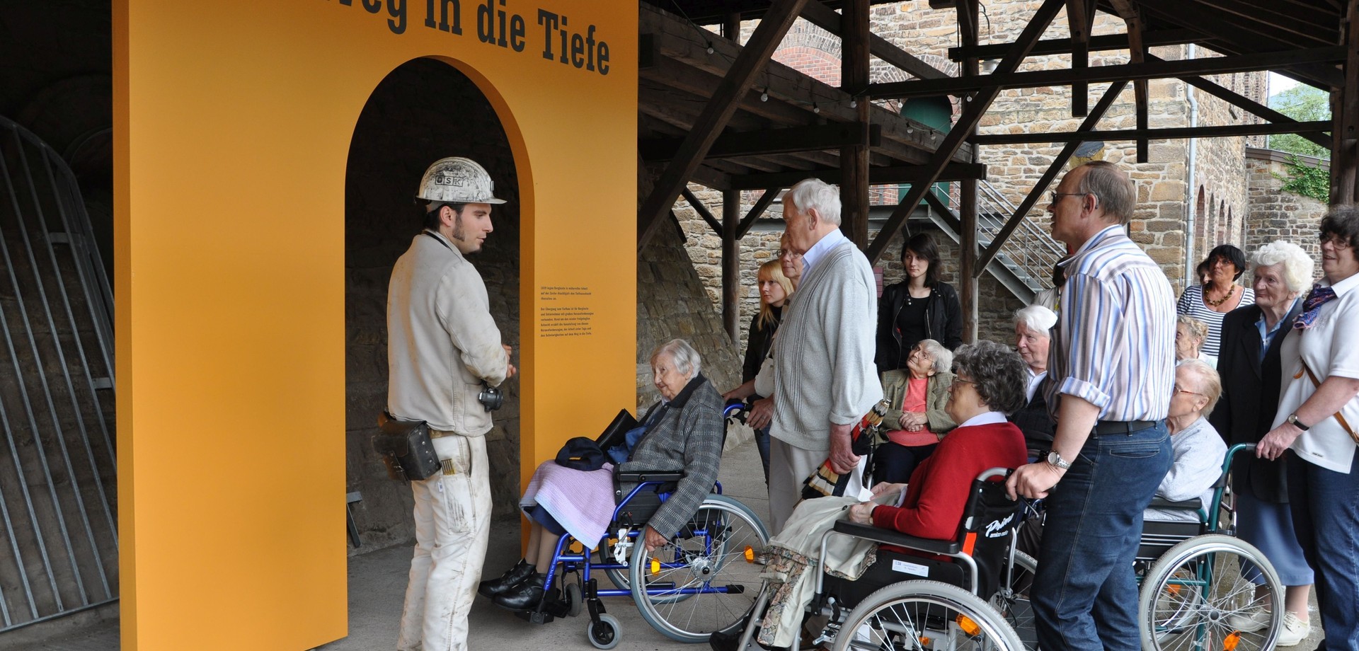 Mehrere Personen im Rollstuhl, stehende Personen und ein Museumsführer haben sich um den Eingang des Bergwerks der Zeche Nachtigall versammelt.