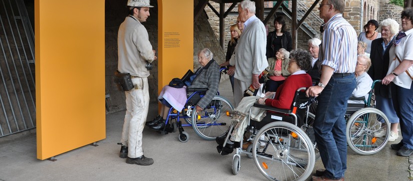 Mehrere Personen im Rollstuhl, stehende Personen und ein Museumsführer haben sich um den Eingang des Bergwerks der Zeche Nachtigall versammelt.