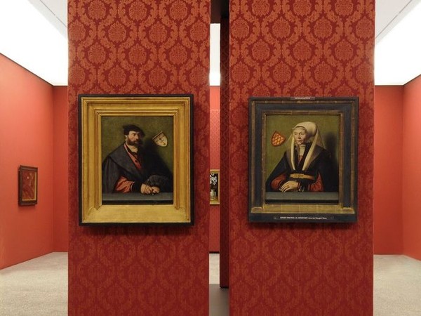 Zwei Gemälde hängen an einer roten Wand in einem Ausstellungsraum.