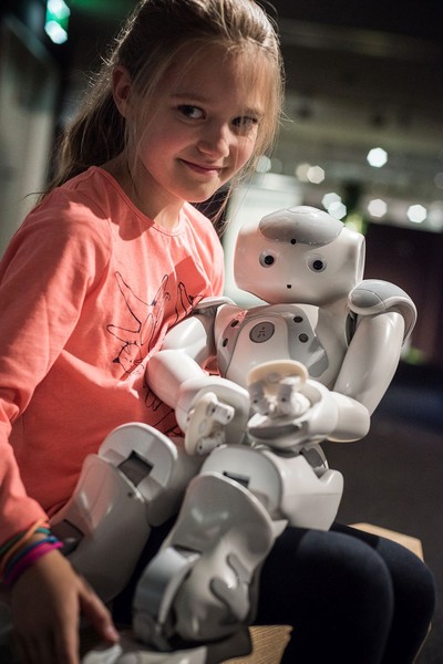 Ein Mädchen guckt lächelnd in die Kamera. Sie hält einen kindlich aussehenden Roboter im Arm.