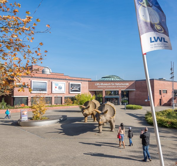 Museumsvorplatz mit Fahnenmast im Vordergrund. In der Bildmitte zwei Dinosaurierfiguren, Menschen, Pflanzen. Im Hintergrund das Museumsgebäude.