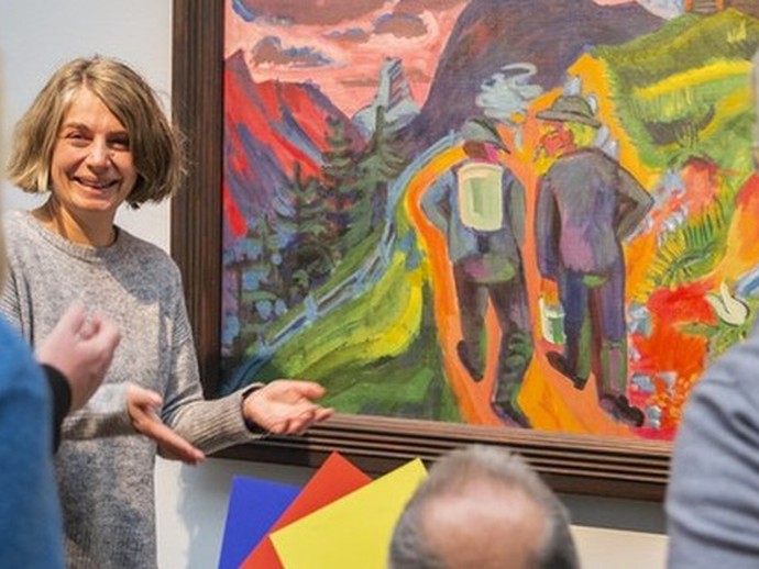 Drei Personen betrachten ein farbenfrohes Gemälde mit zwei Wanderern in einer Berglandschaft in einem Museum. Eine weitere Frau erklärt das Bild.