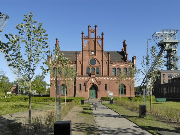 Rotes Backsteingebäude mit gotischen Elementen, flankiert von zwei großen Fördertürmen, umgeben von Bäumen und Grünflächen.