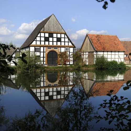 Hinter einem Teich mit belaubten Bäumen sieht man zwei Fachwerkhäuser mit weiß verputzten Gefachen zwischen den Fachwerkbalken aus Holz.