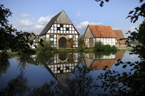 Hinter einem Teich mit belaubten Bäumen sieht man zwei Fachwerkhäuser mit weiß verputzten Gefachen zwischen den Fachwerkbalken aus Holz.