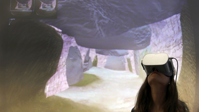 Eine Frau guckt durch eine Virtual Reality Brille. Im Hintergrund sieht man eine virtuelle Nachbildung eines Großsteingrabes.