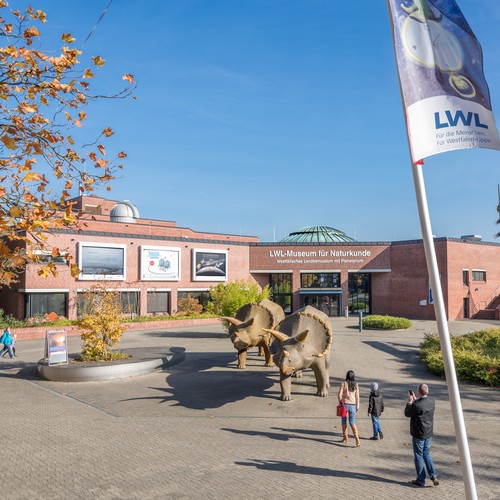 Museumsvorplatz mit Fahnenmast im Vordergrund. In der Bildmitte zwei Dinosaurierfiguren, Menschen, Pflanzen. Im Hintergrund das Museumsgebäude.