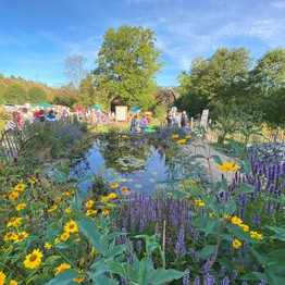 Blick auf einen Teich mit gelben und lila Blumen im Vordergrund und Besuchenden im Hintergrund.