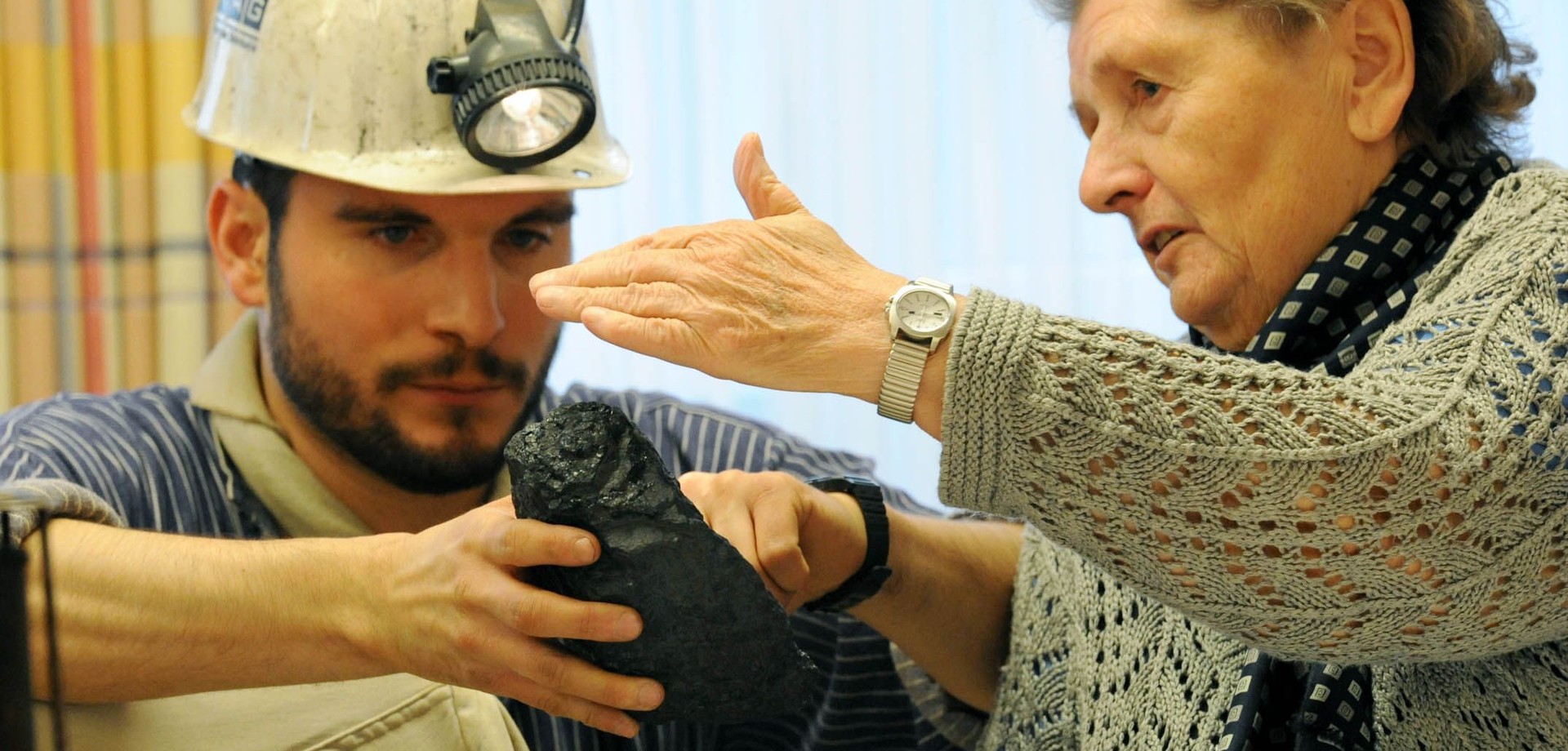 Eine grauhaarige Frau betastet ein Stück Kohle. Ein Museumsführer, verkleidet als Bergarbeiter, hält die Kohle in der Hand.