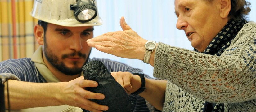 Eine grauhaarige Frau betastet ein Stück Kohle. Ein Museumsführer, verkleidet als Bergarbeiter, hält die Kohle in der Hand.