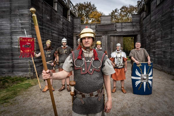 Eine Gruppe von sieben Männern in römischer Soldatenausrüstung vor Holzwänden, vorne ein Mann mit Helm und Speer