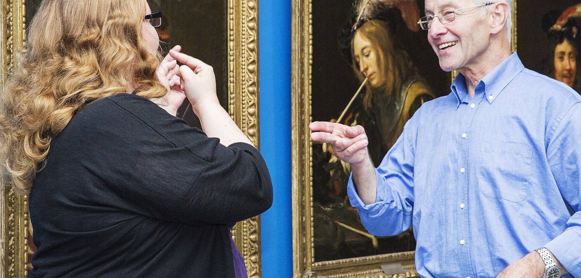 Zwei Menschen stehen in einem Museum vor Gemälden und unterhalten sich in Gebärdensprache.